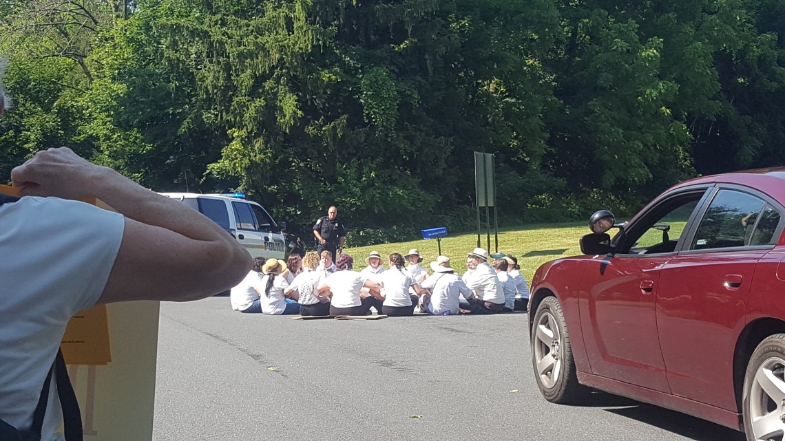 About 17 people sit in a circle in a road, with a police officer standing and looking at them