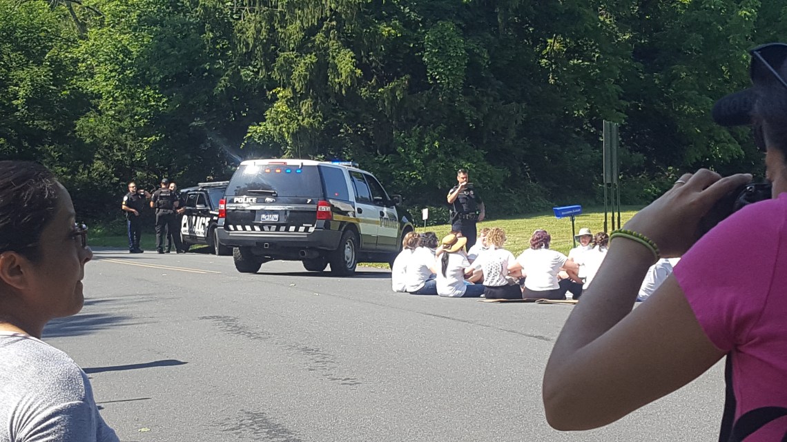 A police officer with a camera watches a group of people seated in a road, and several other officers stand nearby