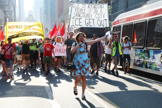 Protesters march in the street, carrying a banner that reads "ICE is rounding people up in a sanctuary city. ICE GET OUT." An activist leads the march with a megaphone.