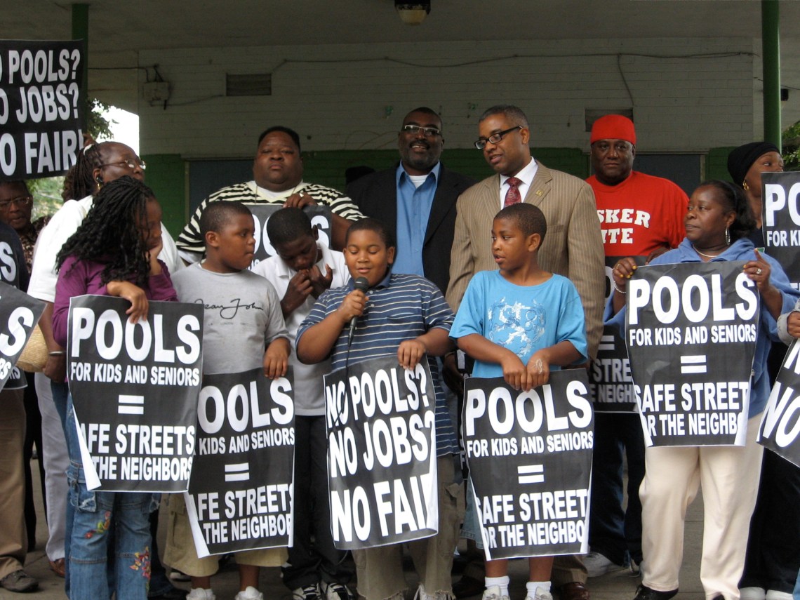 Several children and adults stand holding signs that read "Pools for kids and seniors = safe street for the neighbors" and "no pools? no jobs? no fair!"
