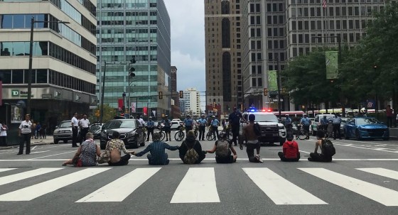 Eight people sit or kneel across an intersection with their hands linked, as police on bicycles and in police cars face them.