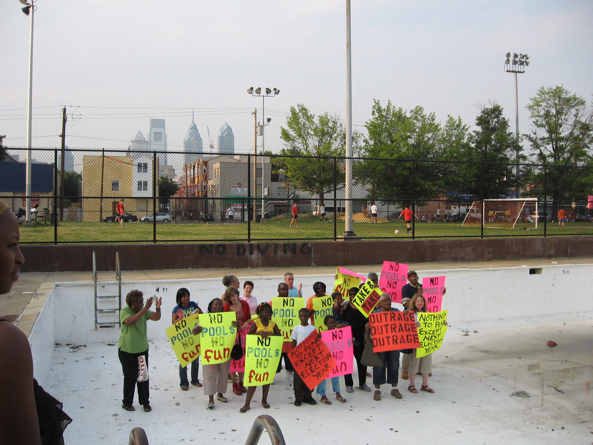 About a dozen children stand in an empty swimming pool holding signs that read "no pools, no fun."