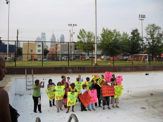 About a dozen children stand in an empty swimming pool holding signs that read "no pools, no fun."