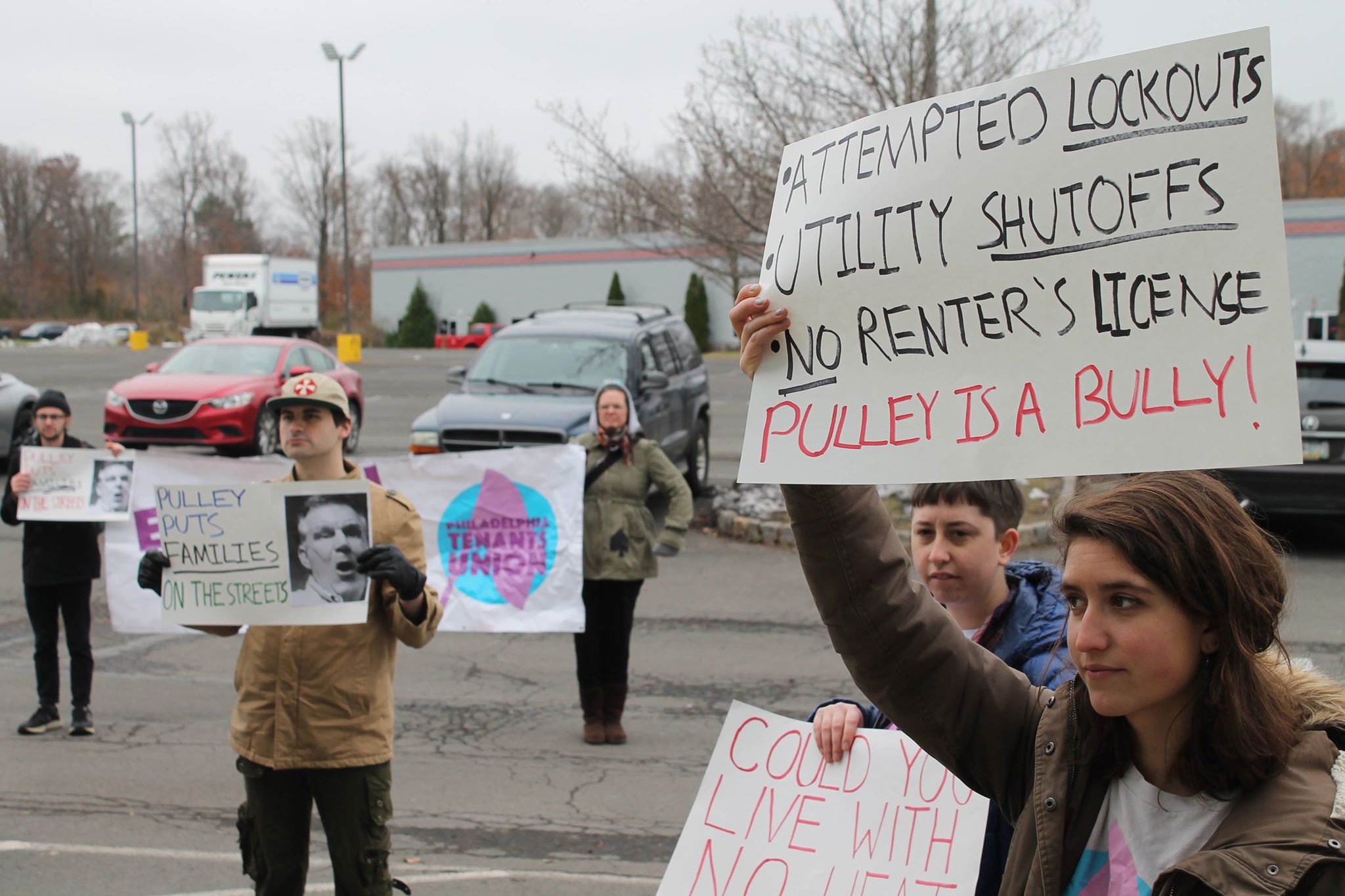 Several activists stand in a parking lot. A sign reads "Attempted lockouts, utility shutoffs, no renter's license, Pulley is a bully!"